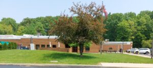 green field of grass with a tree in the center and tan brick building on the left.