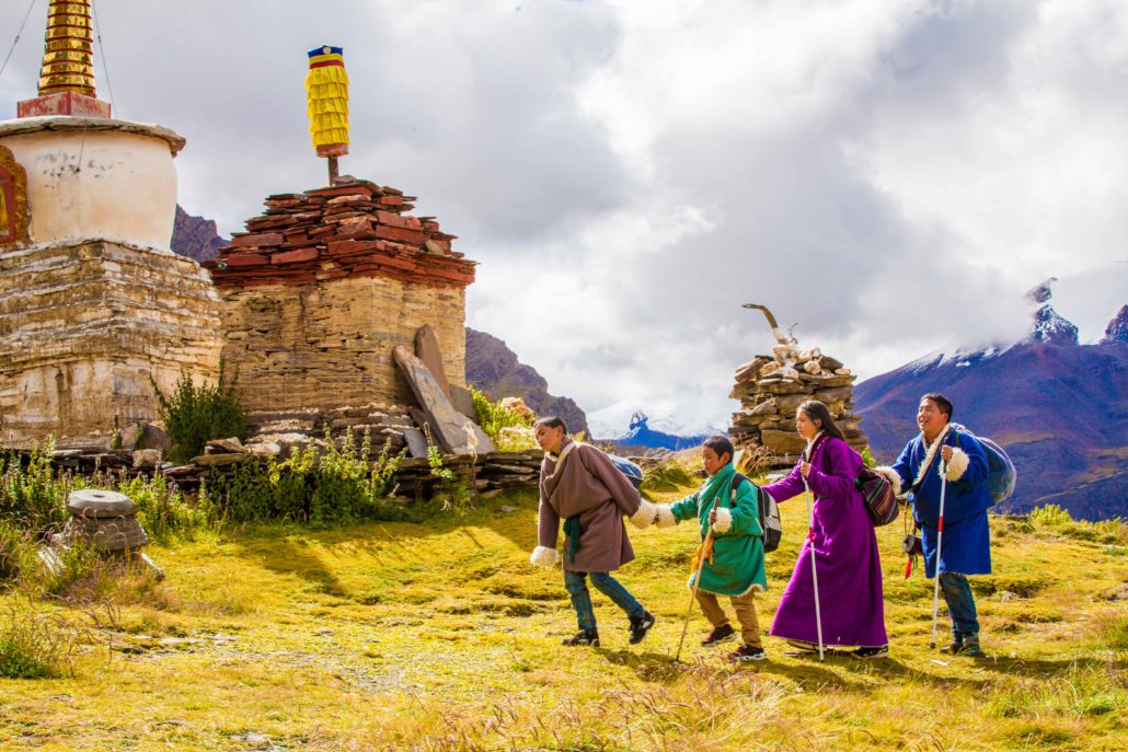 Four small children walk by a Tibetan temple, guiding each other by placing their hands on each others shoulders. Mountains in the background.