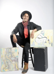 Artist Bryan Moss sits on a brown stool. He has brown skin and is wearing a black suit with a red scarf. His hand rests on some artwork. 