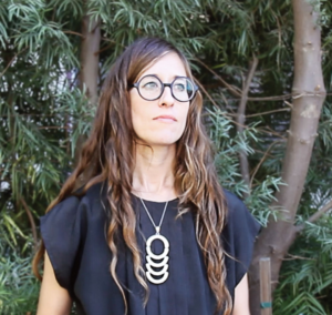 An image of Allison O'Daniel, a white woman with long, curly brown hair and black, round glasses. She wears a black shirt and an elaborate silver necklace. Behind her is some green foliage and tree trunks.