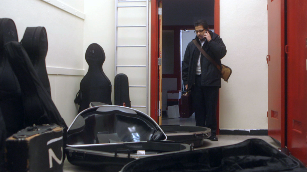 A man stands in the open door of a small room with white walls. He holds a black instrument case in one hand and a cell phone to his ear in the other. Before him, on the floor are a few large empty instrument cases. The top of the cases are open.