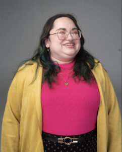 Portrait of Rebecca Gonzalez-Bartoli. The background is grey. Rebecca has long dark hair and is wearing glasses, earrings, a heart shaped necklace, a pink turtleneck with a golden sweater and black pants with a silver belt.