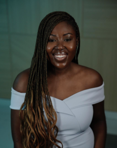 Portrait of Cynthia Amoah smiling with hair over her right shoulder. She is wearing a white dress.