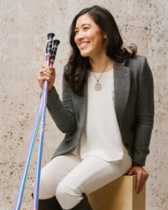 A young asian woman with long brown hair, wearing white pants and top with a gray suit jacket, seated on a wooden cube, holding light blue and lilach ski poles.