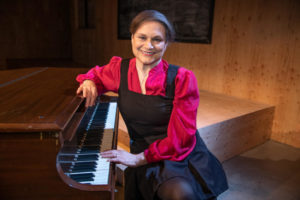 A woman with short brown hair sits at the piano, smiling.