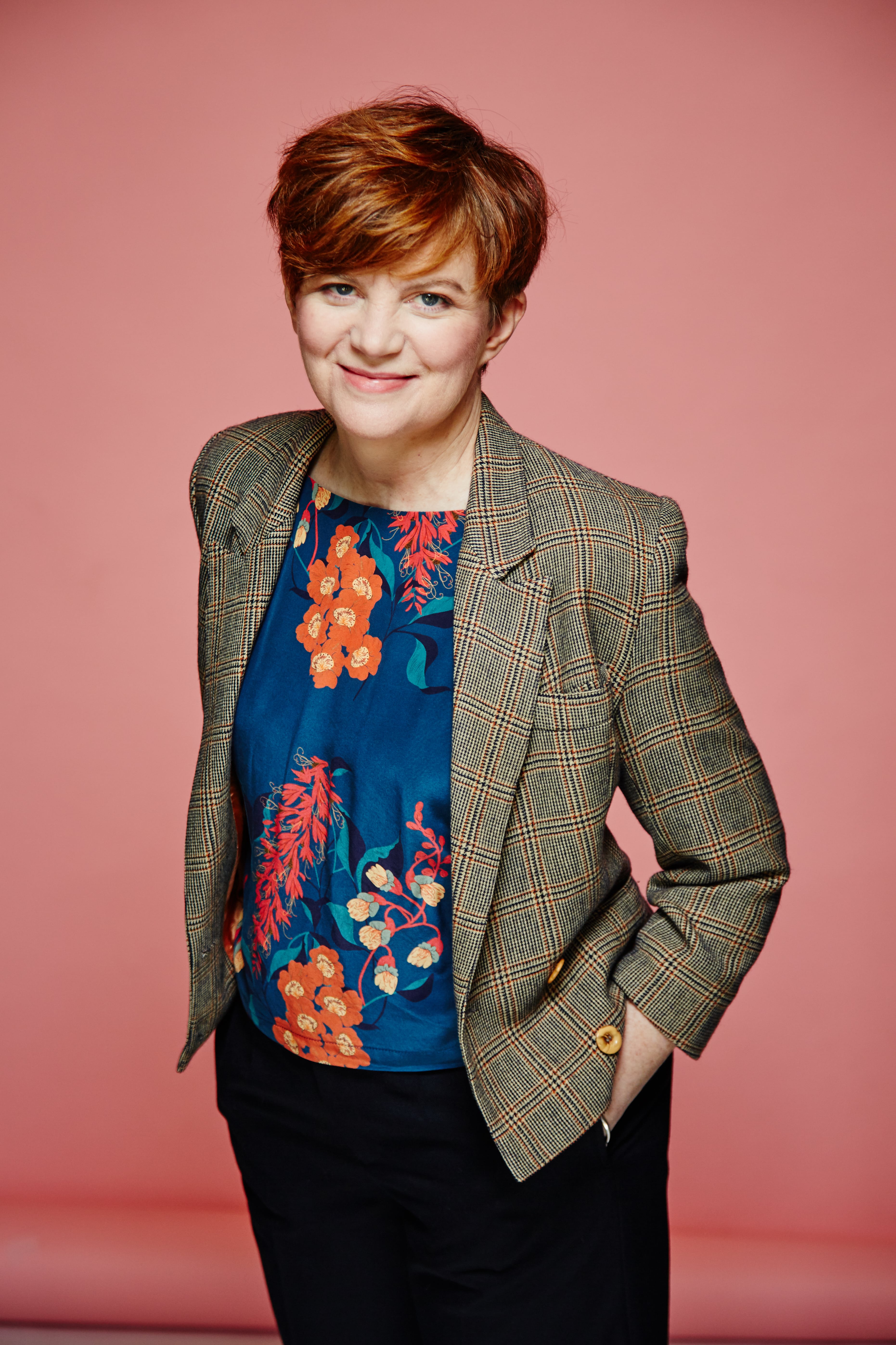A short-cut redhead woman wearing a floral blue and orange shirt, with a grey blazer on top, hands in her pockets, on a pink background