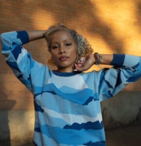 A young woman with blond curly hair, wearing a blue sweater, holding her arms above her shoulders,