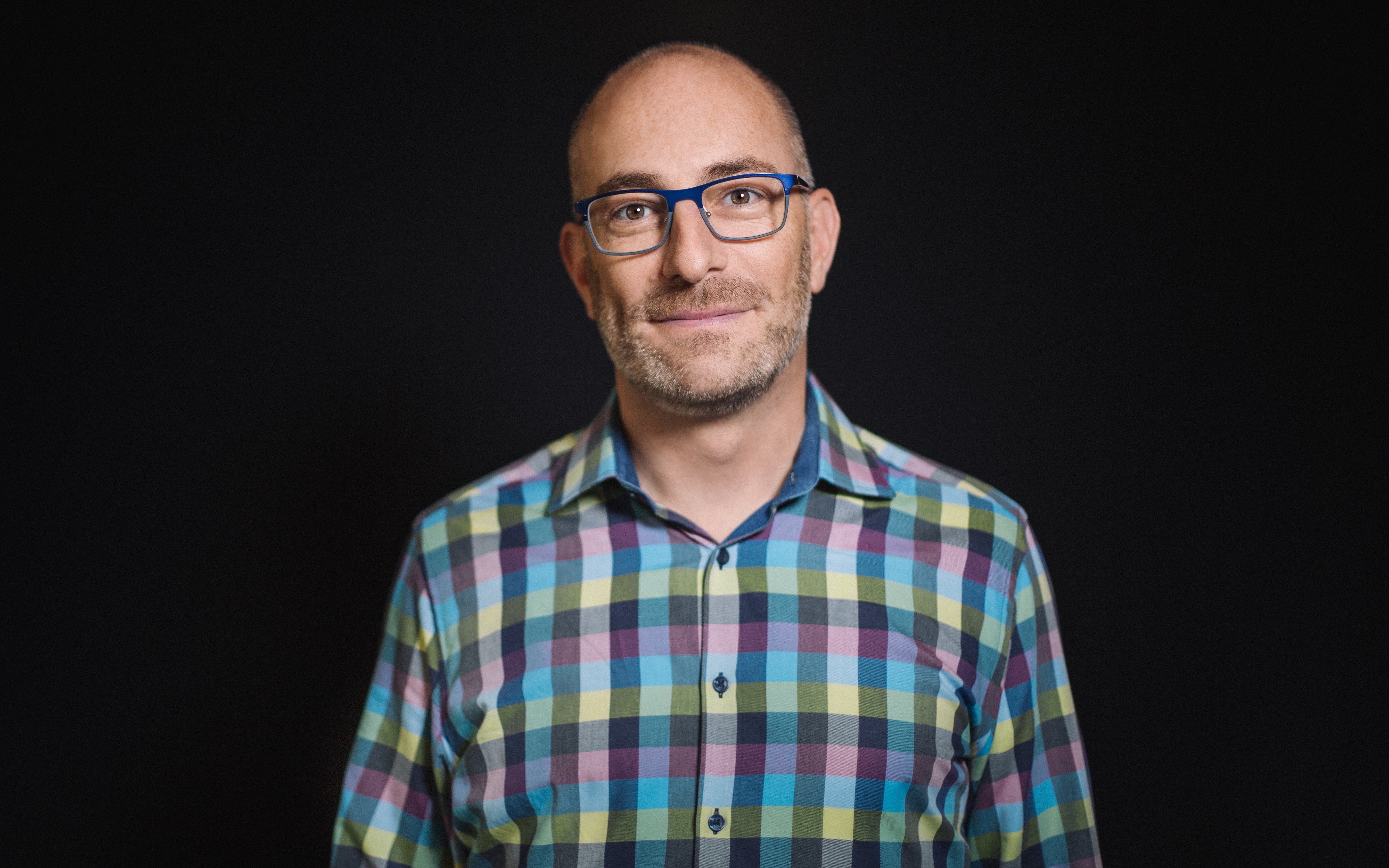 Headshot - Andrew Simon. A man with a colorfull plaid shirt, blue framed glasses, and a kind smile