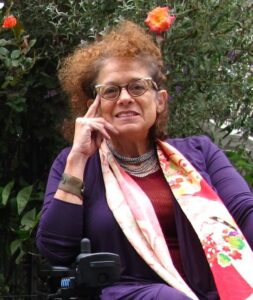 An image of Simi Linton - a woman with curly light brown hair, wearing glasses, a floral pattern scarf and a purple sweater, leaning her head on her right hand