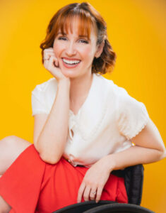 young woman seated in a wheelchair, wearing a red skirt, white shirt, leaning her head on her right palm, smiling. On a bright yellow background
