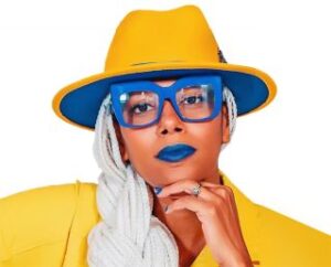 A woman dressed in wide rim yellow hat and a yellow suit, long braided hair, and blue glasses and lipstick.