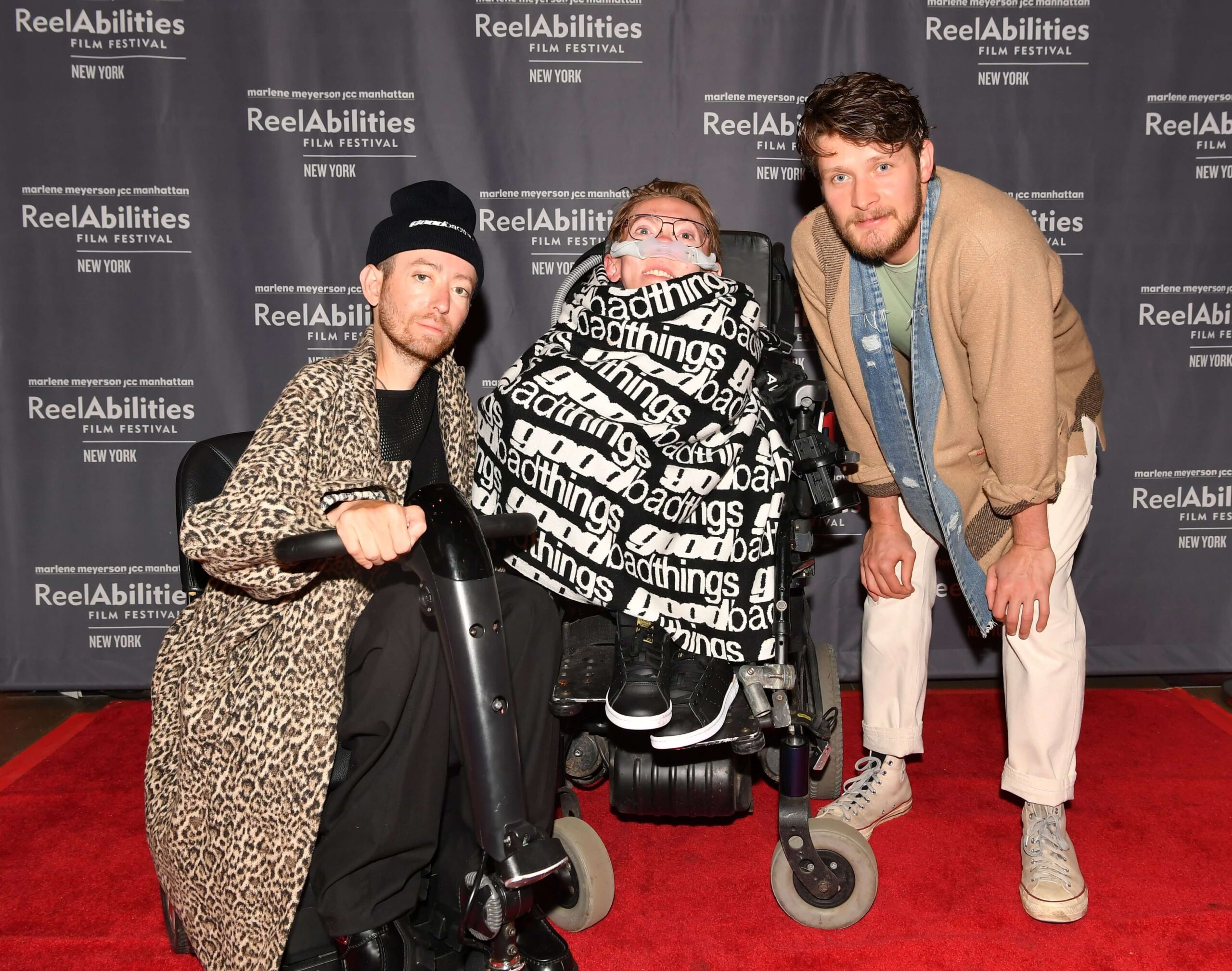 Actor Danny Kurtzman with a beanie cap and leopard pattern long coat, seating in a mobility device, Executive Producer Steve Way covered with a blanket with a text pattern saying Good Bad Things in white letters over black background, Actor Brett Dier in a brown jacket and white pants leaning forward. All three are lined up on a red carpet, with a banner carrying the ReelAbilities Film Festival logo behind them.
