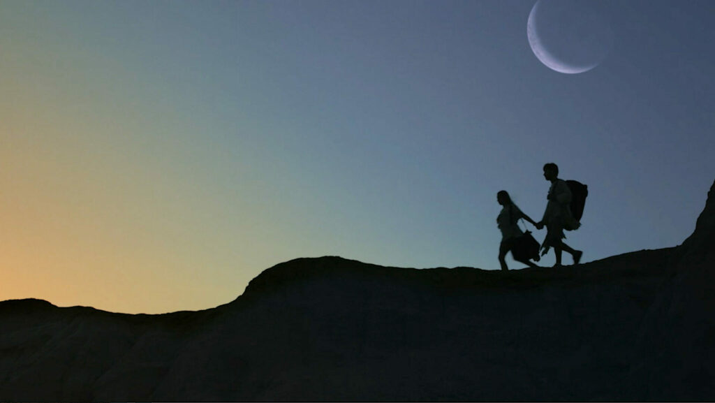 Shadows of two people appear on a mountain under a crescent moon and dusky sky.