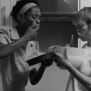 Two women hold a bowl in the kitchen and eat from it, licking their fingers.