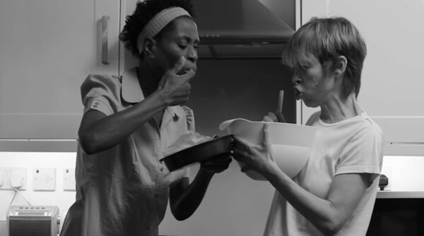 Two women hold a bowl in the kitchen and eat from it, licking their fingers.
