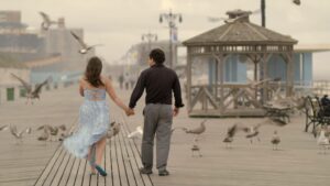 A couple holds hands on a boardwalk in front of a wooden gazebo, surrounded by birds and a hazy sky.
