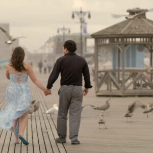 A couple holds hands on a boardwalk in front of a wooden gazebo, surrounded by birds and a hazy sky.
