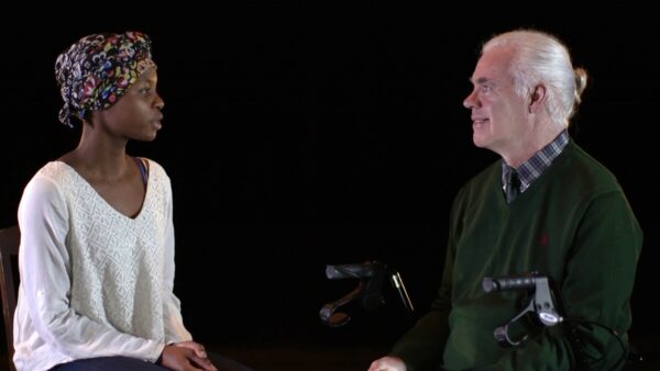 A silver haired white man sits in front of a black background, looking at a young black woman in white with a floral headwrap.