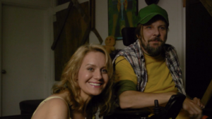 A man in a green cap and yellow shirt sits in a wheelchair next to a blonde woman who smiles at the camera.
