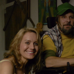 A man in a green cap and yellow shirt sits in a wheelchair next to a blonde woman who smiles at the camera.