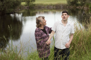 A young man in a white shirt stands next to an older woman in purple, who looks at him and touches his shoulder, smiling. The two stand in front of a pond with grass in the foreground of the shot.