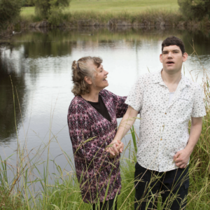 A young man in a white shirt stands next to an older woman in purple, who looks at him and touches his shoulder, smiling. The two stand in front of a pond with grass in the foreground of the shot.