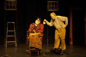 Two older white men appear on a stage; one sits in a wooden wheelchair, the other stands over him with a metal clock.