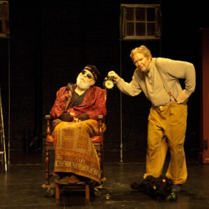 Two older white men appear on a stage; one sits in a wooden wheelchair, the other stands over him with a metal clock.