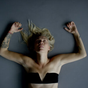 A blonde woman lies on her back on a dance studio floor with her arms up by her head as she looks at the ceiling.