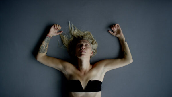 A blonde woman lies on her back on a dance studio floor with her arms up by her head as she looks at the ceiling.