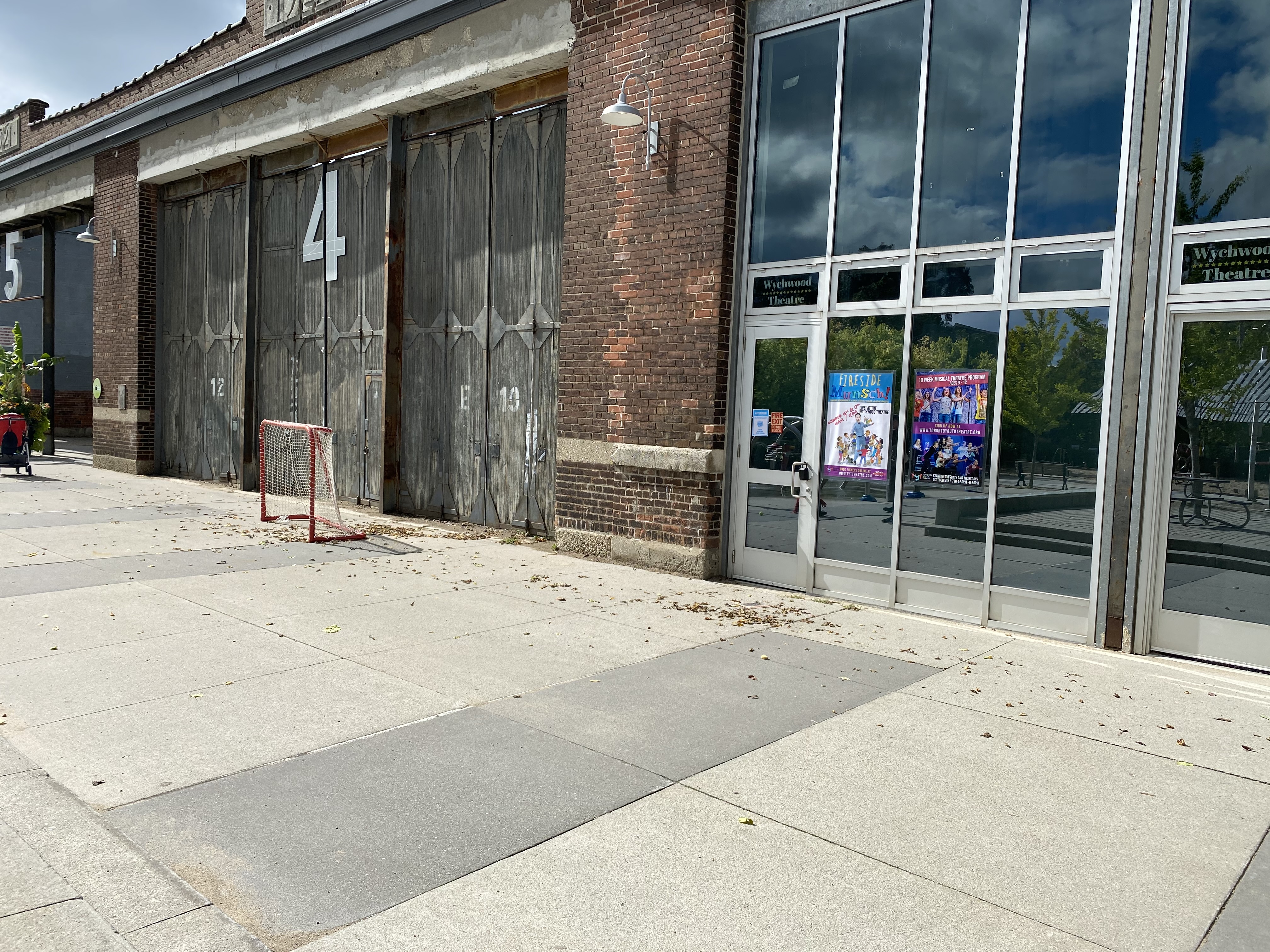 This is the area where Dis/Play will be projected. The ground features a smooth, paved surface, with grass and trees in the background, and the building landmark is the door to Barn 4, on the side of ArtScape Wychwood Barns.
