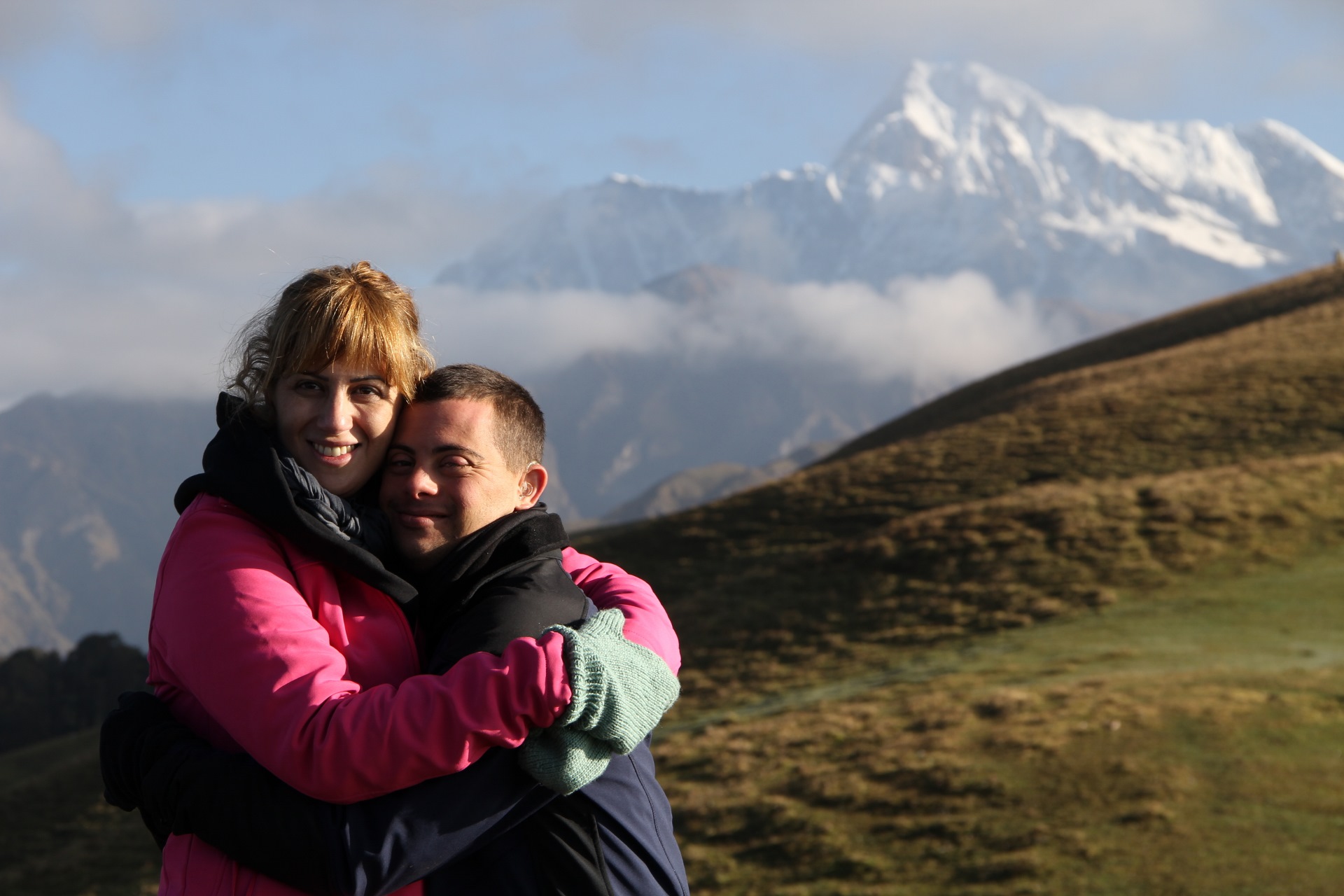 A man and woman hug in a green field with mountains behind them