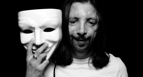 a man with long black hair, with some deformation in his facial structure, is holding a white theater mask to the side of his face