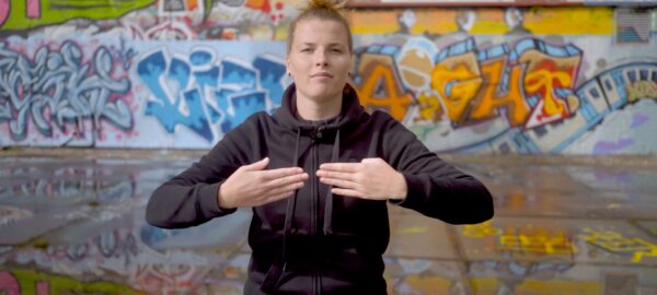 a young woman standing on the background of a graffiti covered wall, with her arms in front of her chest in a signing gesture.