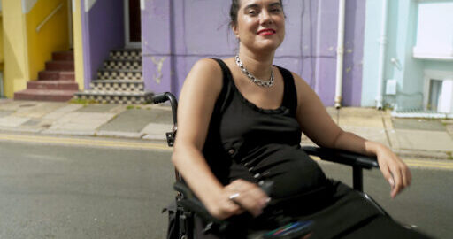 a young woman seated in a motorised wheelchair smiling, on the background of colourful old buildings