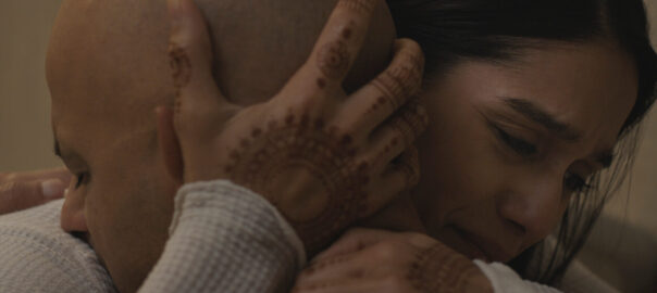 A dark-haired woman with an expression of deep concern embraces a man and clasps his head with a henna-decorated hand.