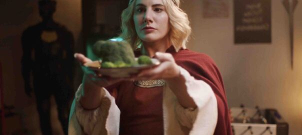 A blonde woman wearing a medieval outfit with draping white sleeves, a crown and a red cape presents a plate of broccoli.