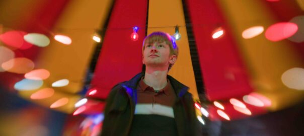 A blonde boy gazes at the colorful string lights surrounding him, with a striped red and yellow backdrop.