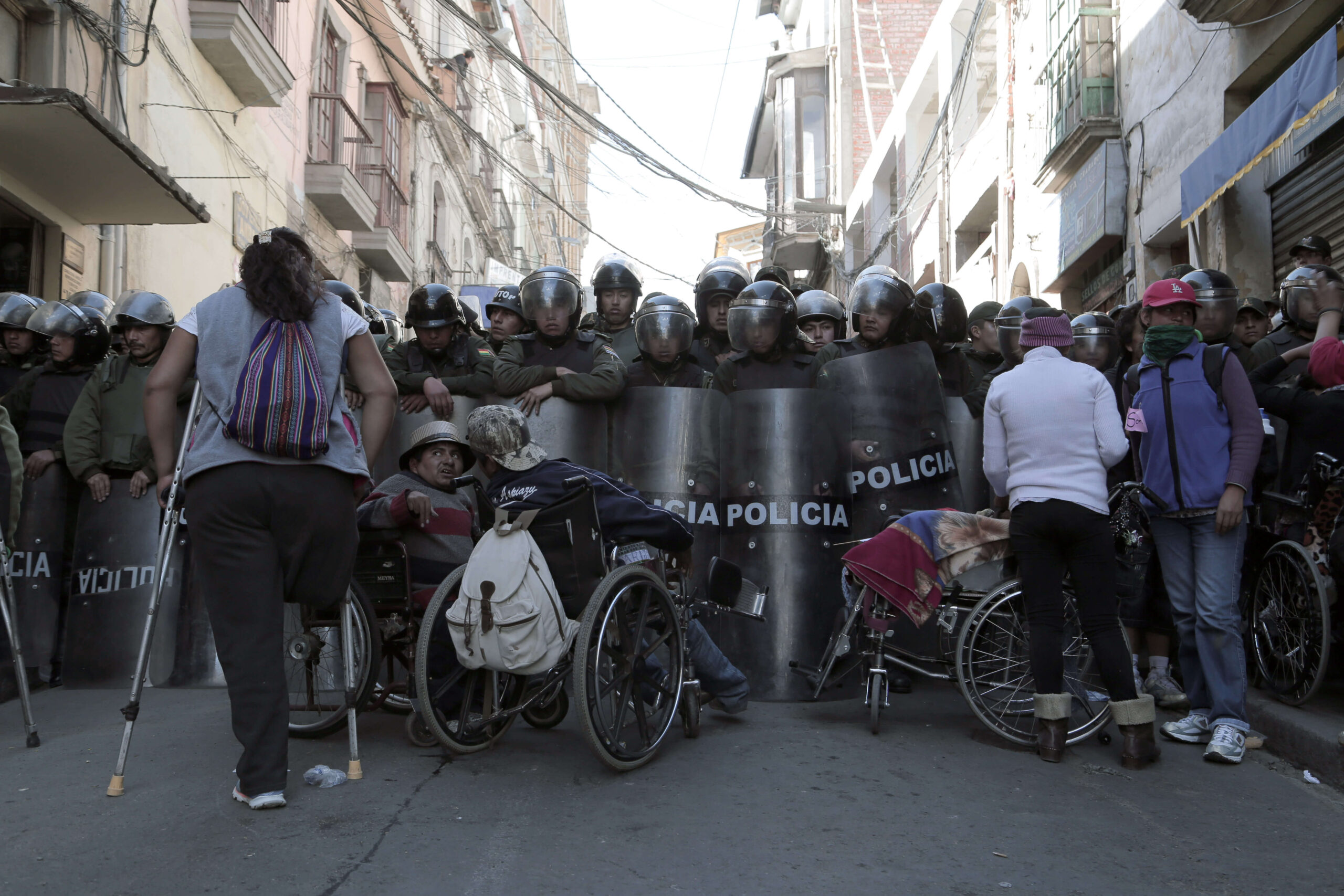 A group of police officers in full gear stand with shields in front of four activists with various disabilities.