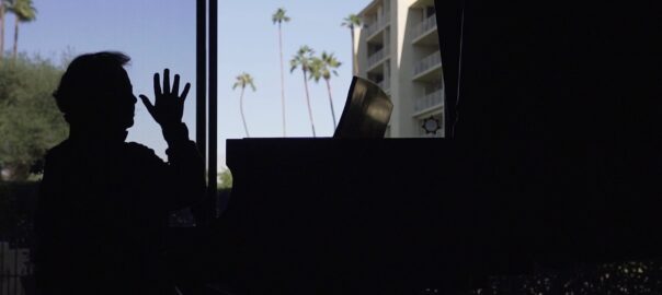 A man in shadow is backlit by a window; he holds his hand in the air over a piano.