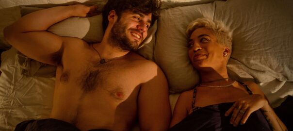 A black woman with short blonde smirks at the man with dark hair lying beside her, grinning back.
