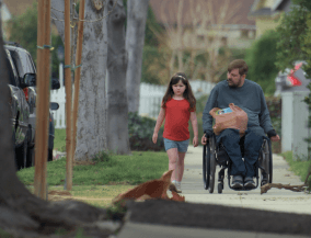 A man in a wheelchair looks at a girl accompanying him on a sidewalk.