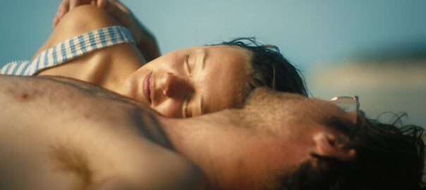 A man looks away from the camera at the woman lying beside him with her eyes closed at the beach, with the sun beating down on them with a hazy blue backdrop.