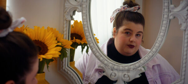 A white woman with dark red lipstick and dark brown hair pulled into a bun with a pink bow peers into a white framed mirror, showing her reflection and that of a sunflower beside her. She has a complex expression with a small but sad smile.