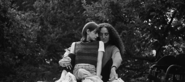 Black and white: A black woman with long hair embraces a white woman with cropped hair from behind as they sit on a picnic blanket on grass, surrounded by trees.
