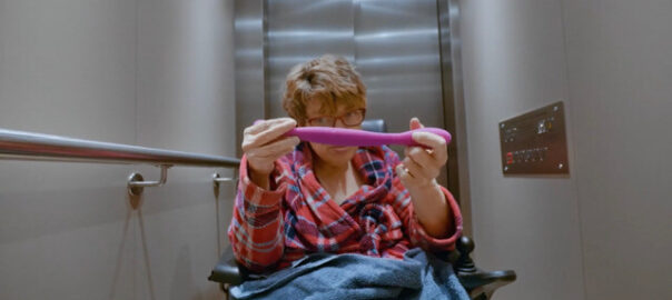 A woman in a wheelchair sits in front of an elevator, holding a pink vibrator in front of her.