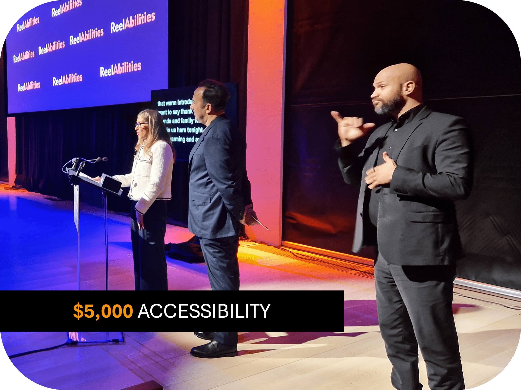 A woman speaking at a podium, a man in a suit stands proudly at her side, in the background is a screen with captions and next to them an ASL interpreter. Text reads: $5,000 Accessibility