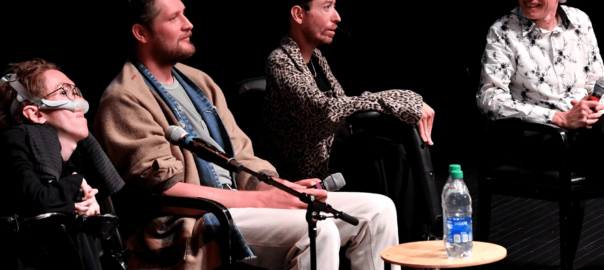 4 men on stage, the moderator is smiling at the panelists - two of them using a wheelchair.